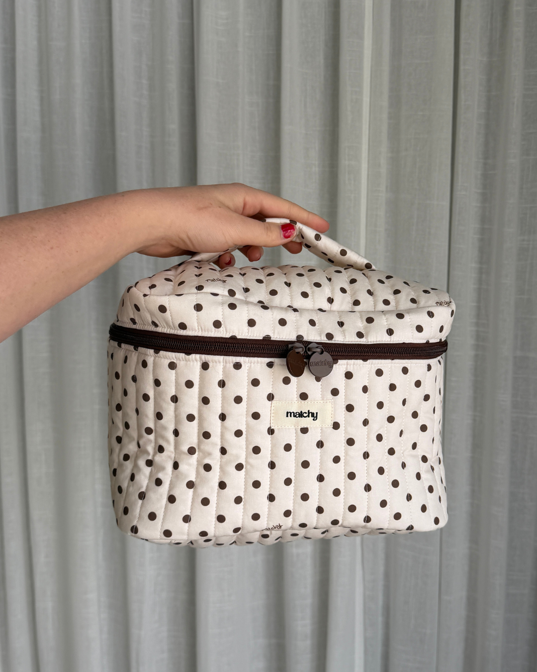 Hand holding a cream quilted train case with brown polka dots, a brown zip and 'matchy' label by its top handle. Matchy Ken Train Case, a quilted makeup and toiletry case.
