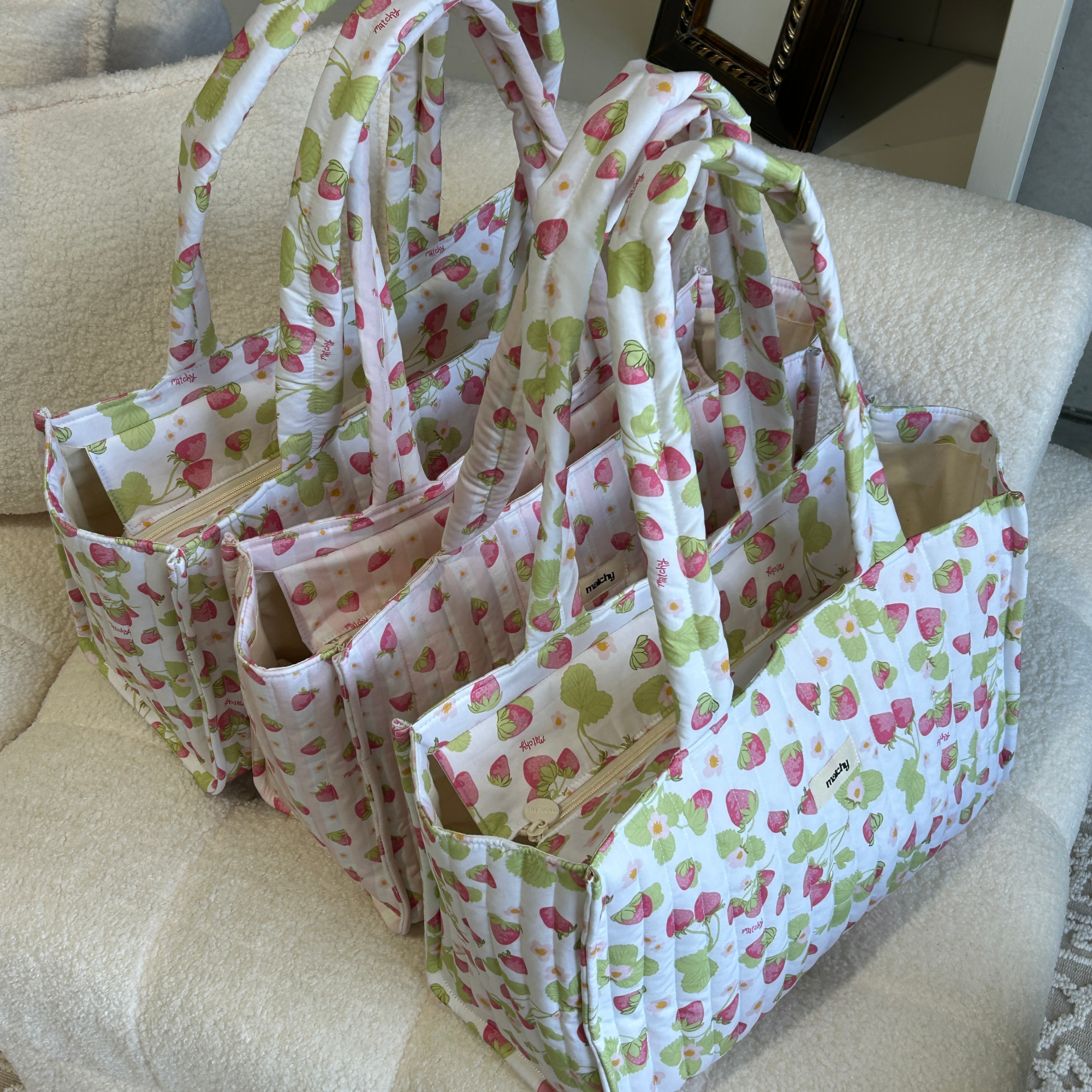 Several strawberry print quilted tote bags lined up on a cream boucle armchair. Matchy Sofia Take Me With You Tote.