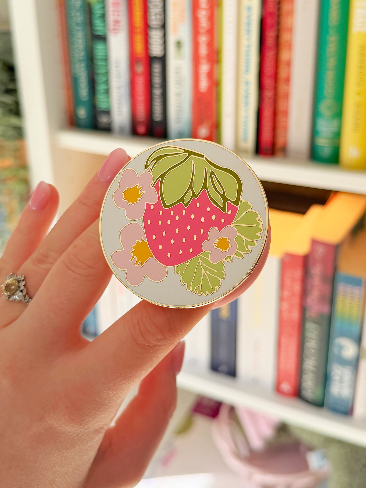 Hand holding a round grip printed with a pink strawberry, green leaves and flowers in front of a bookshelf. Matchy Sofia MagSafe Grip for Kindle and phone.