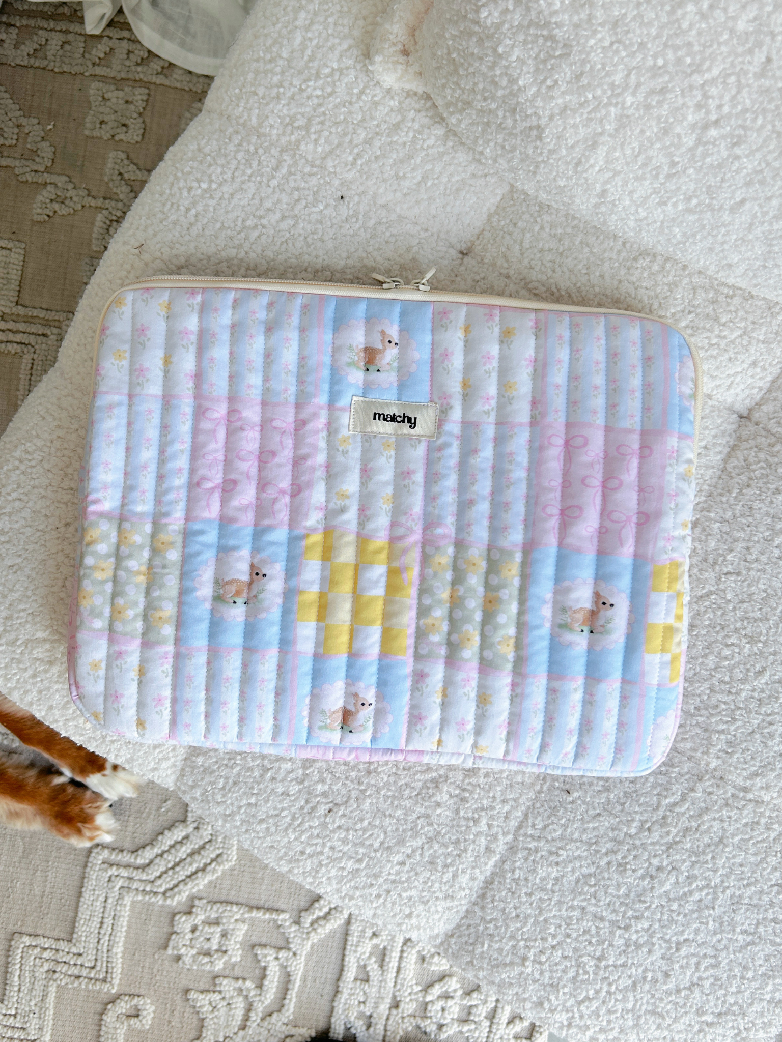 Pastel patchwork quilted laptop sleeve in pink, blue and yellow squares with deer motifs, lying on a cream textured rug. Matchy Patchwork Laptop Sleeve for 13 to 15 inch laptops.