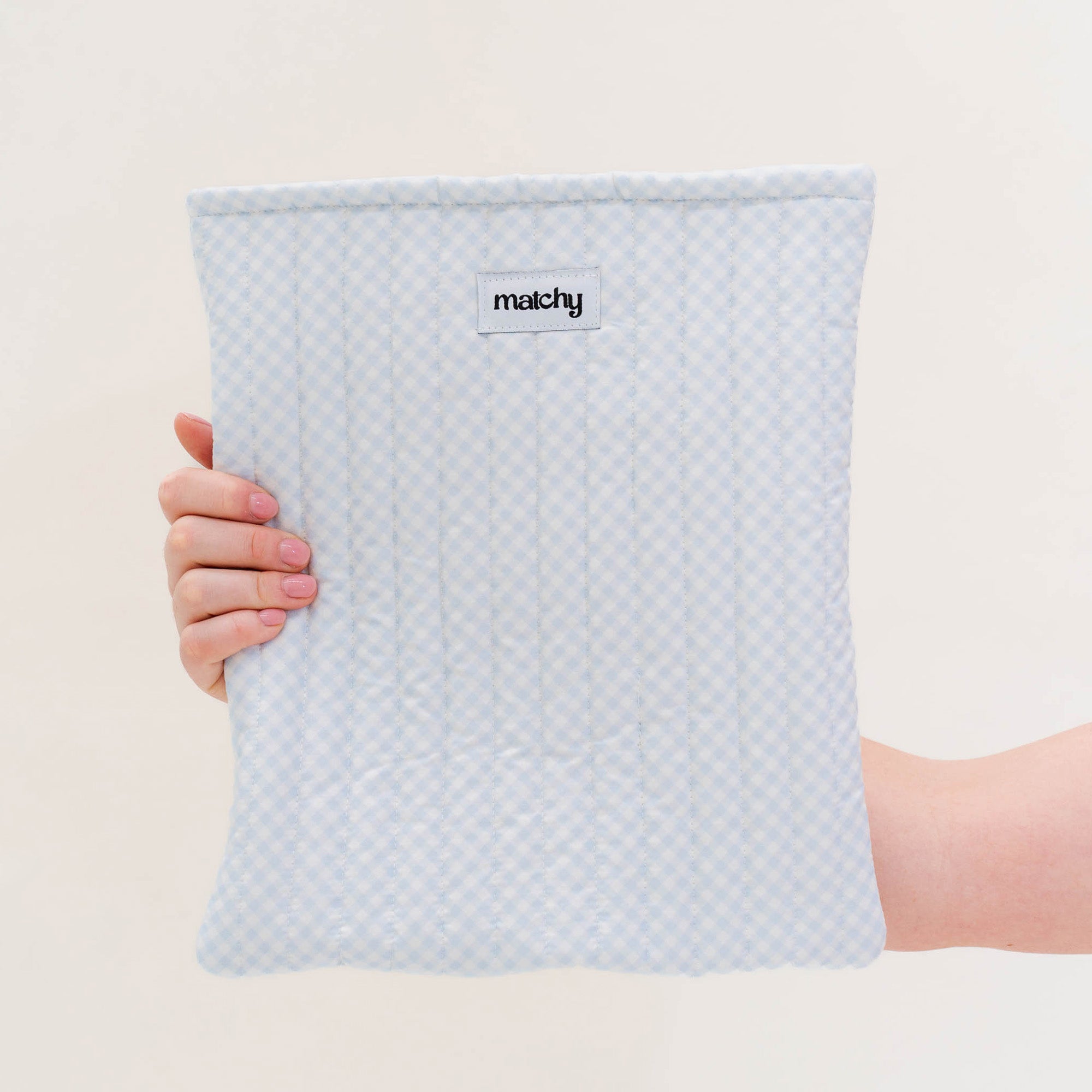 Hand holding a pale blue gingham quilted book sleeve with a 'matchy' label. Matchy Blueberry Gingham Custom Embroidery Book Sleeve, a personalised padded book sleeve.