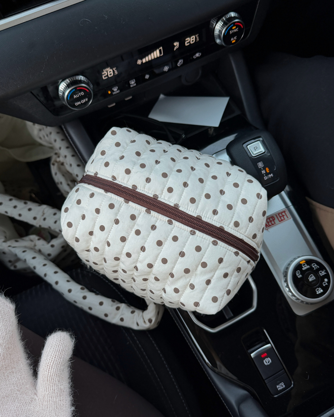 Cream quilted carry-all bag with brown polka dots and a brown zip sitting in a car centre console. Matchy Ken Carry-All Bag, a quilted makeup and toiletry bag.