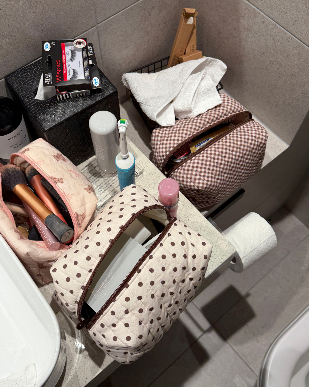 Open cream and brown polka dot carry-all bag on a bathroom bench beside other makeup bags, brushes and skincare. Matchy Ken Carry-All Bag.