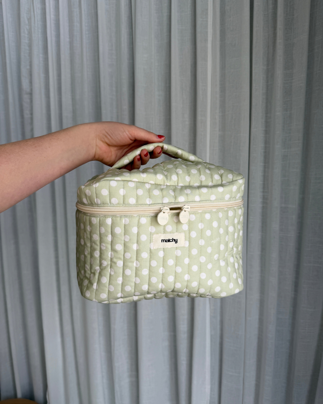 Hand holding a sage green and white polka dot quilted train case by its top handle against a grey curtain. Matchy Poppy Train Case, a quilted makeup and toiletry case.