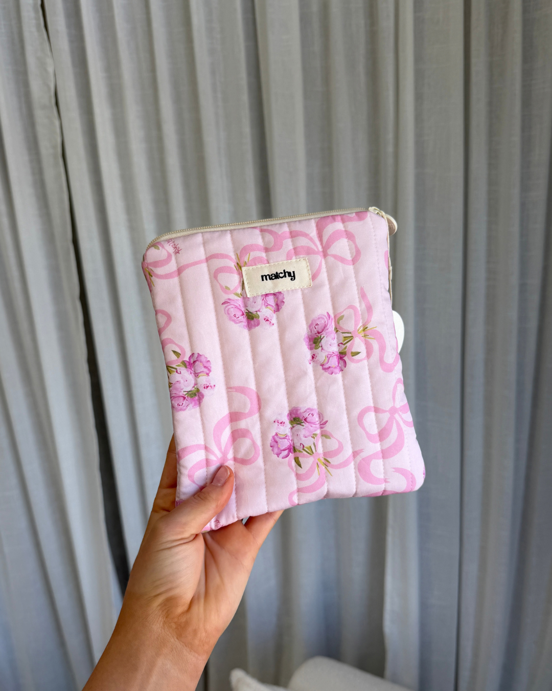 Hand holding a soft pink quilted eReader sleeve printed with pink bows and peonies, with a 'matchy' label. Matchy Evermore Peony eReader Sleeve, a padded Kindle pouch.