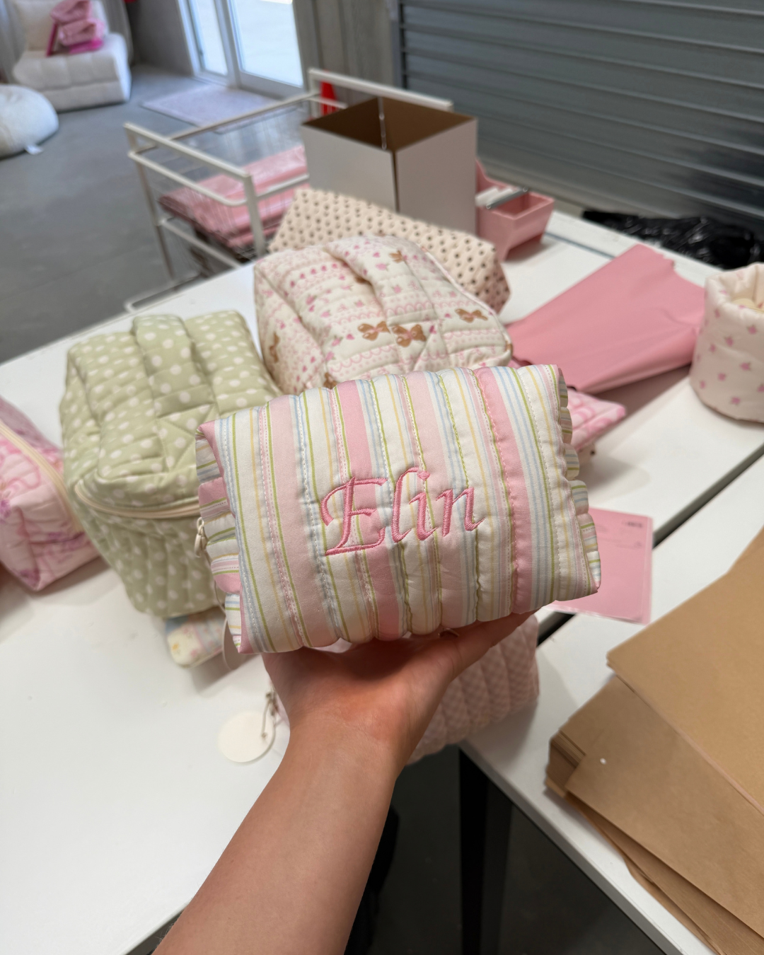 Hand holding a pastel striped carry-all bag embroidered with the name 'Elin' in pink script, in a studio with other bags. Matchy Signature Stripe Custom Embroidery Carry-All Bag.