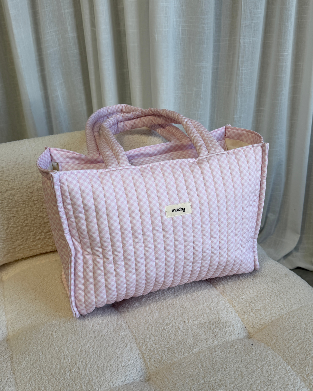 Soft pink gingham quilted tote bag sitting on a cream boucle sofa. Matchy Strawberry Take Me With You Tote, a gingham quilted tote bag.