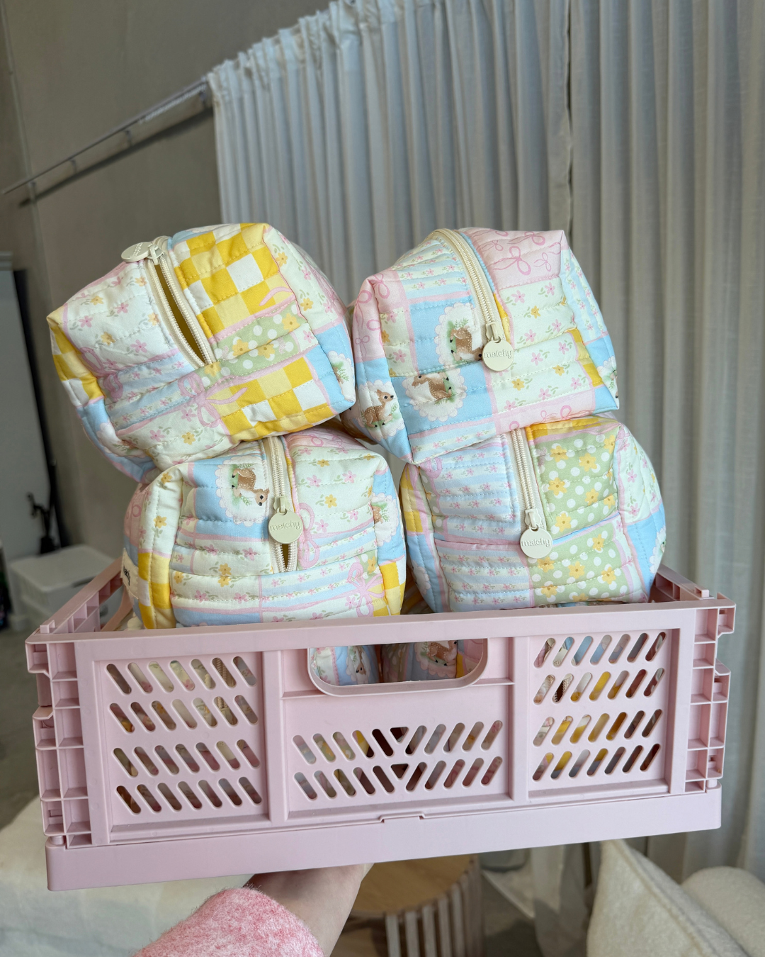 Four pastel patchwork quilted carry-all bags stacked in a pink plastic crate. Matchy Patchwork Carry-All Bag, a quilted makeup and toiletry bag.