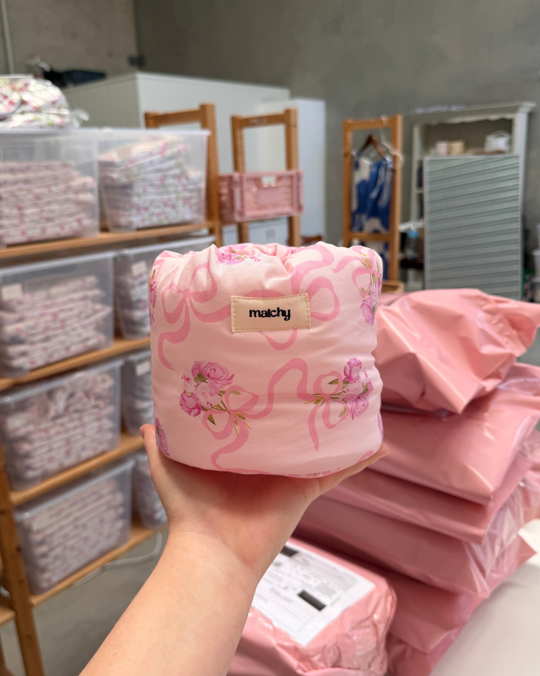 Hand holding a soft pink peony and bow print bubble bucket in the Matchy studio with shelves of stock and pink mailer bags behind. Matchy Evermore Peony Bubble Bucket.