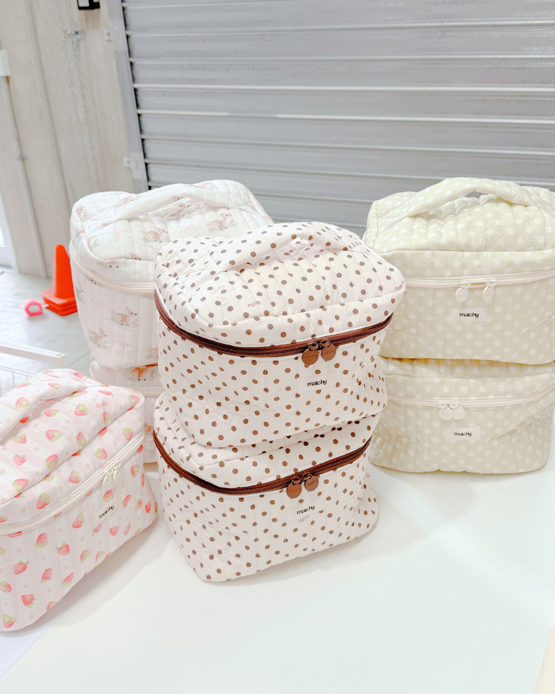 Two cream and brown polka dot train cases stacked on a studio bench beside other quilted cases. Matchy Ken Train Case.