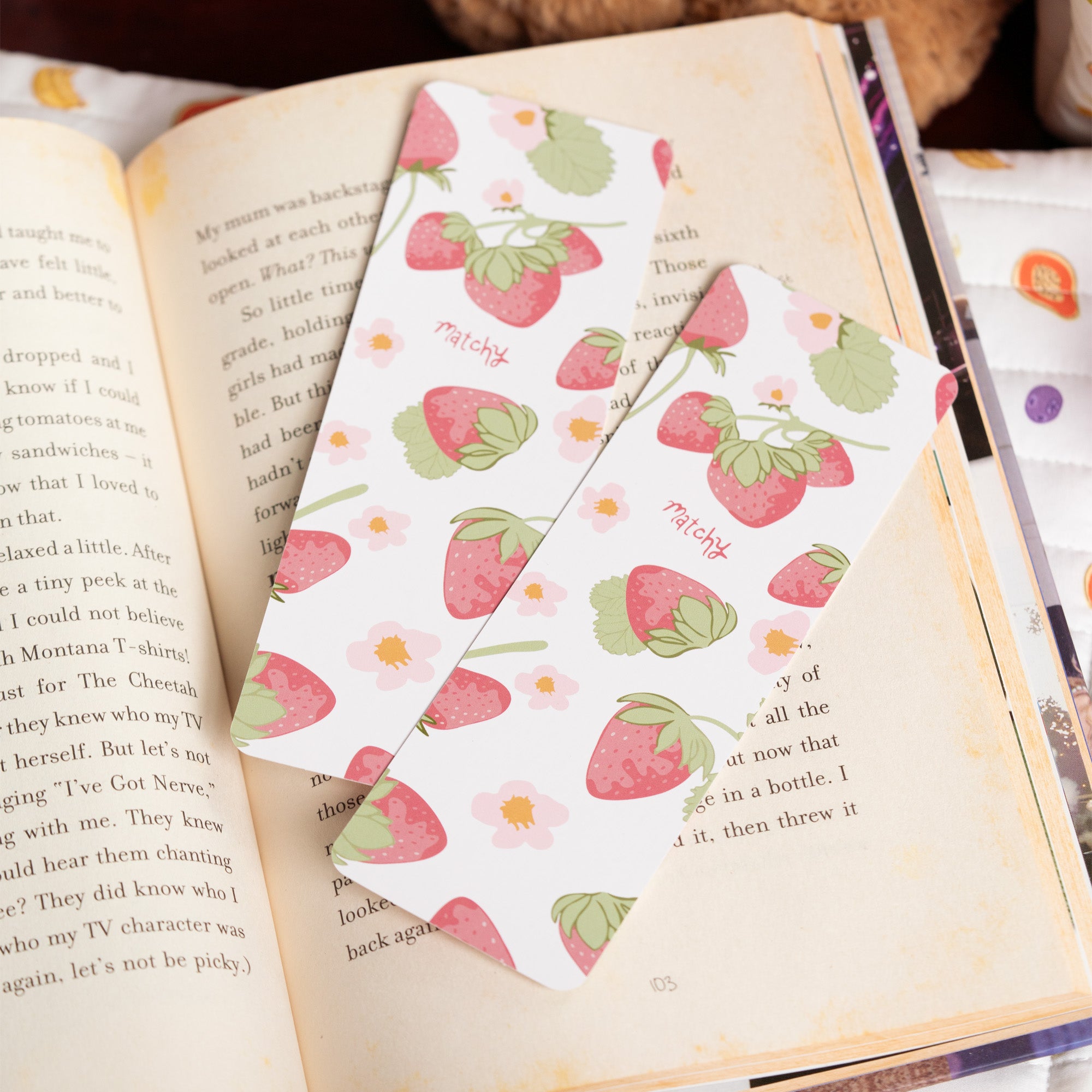 Two cream bookmarks printed with pink strawberries and green leaves resting on an open book. Matchy Sofia Bookmark.