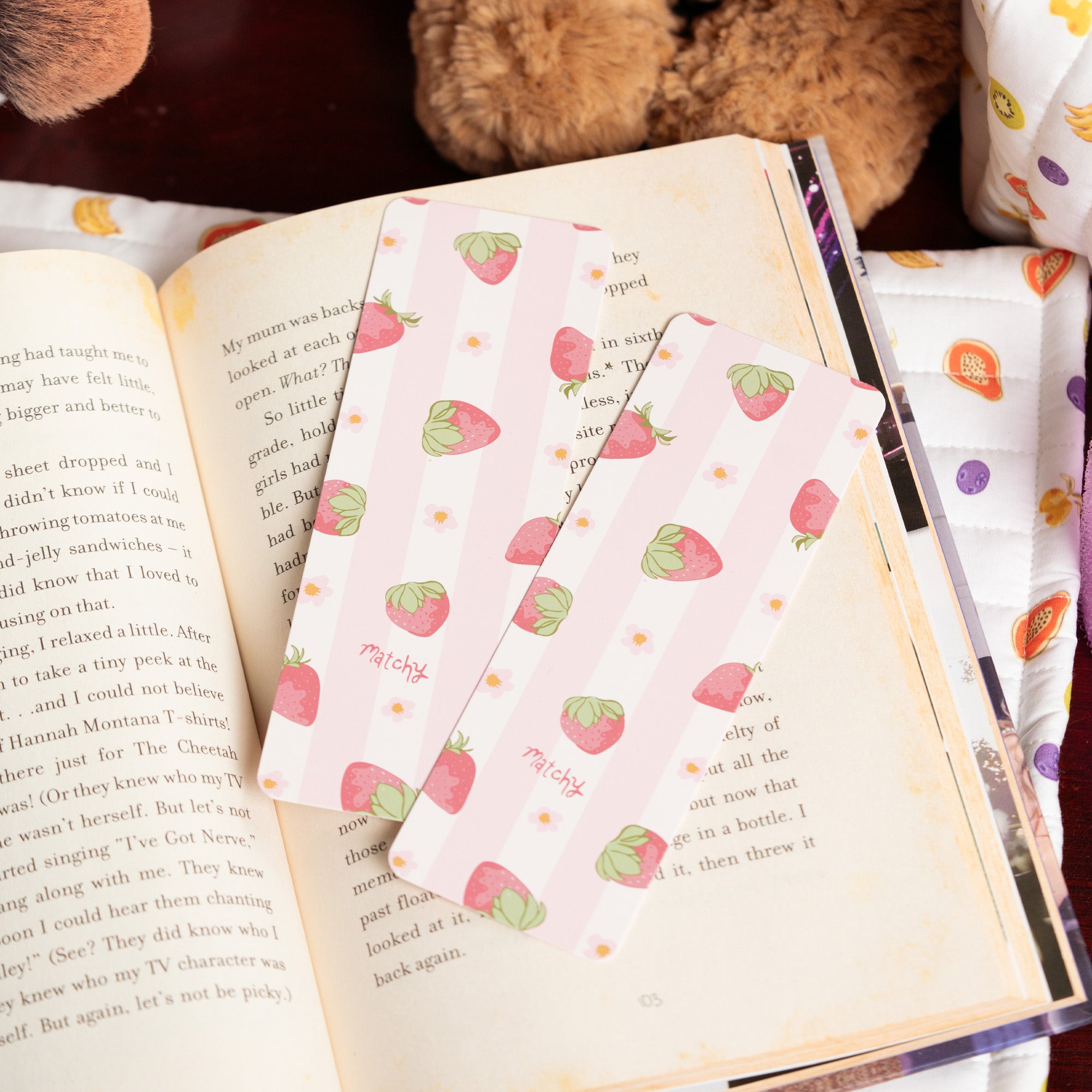 Two pale pink strawberry print bookmarks resting on the pages of an open book. Matchy Sylvie Bookmark.