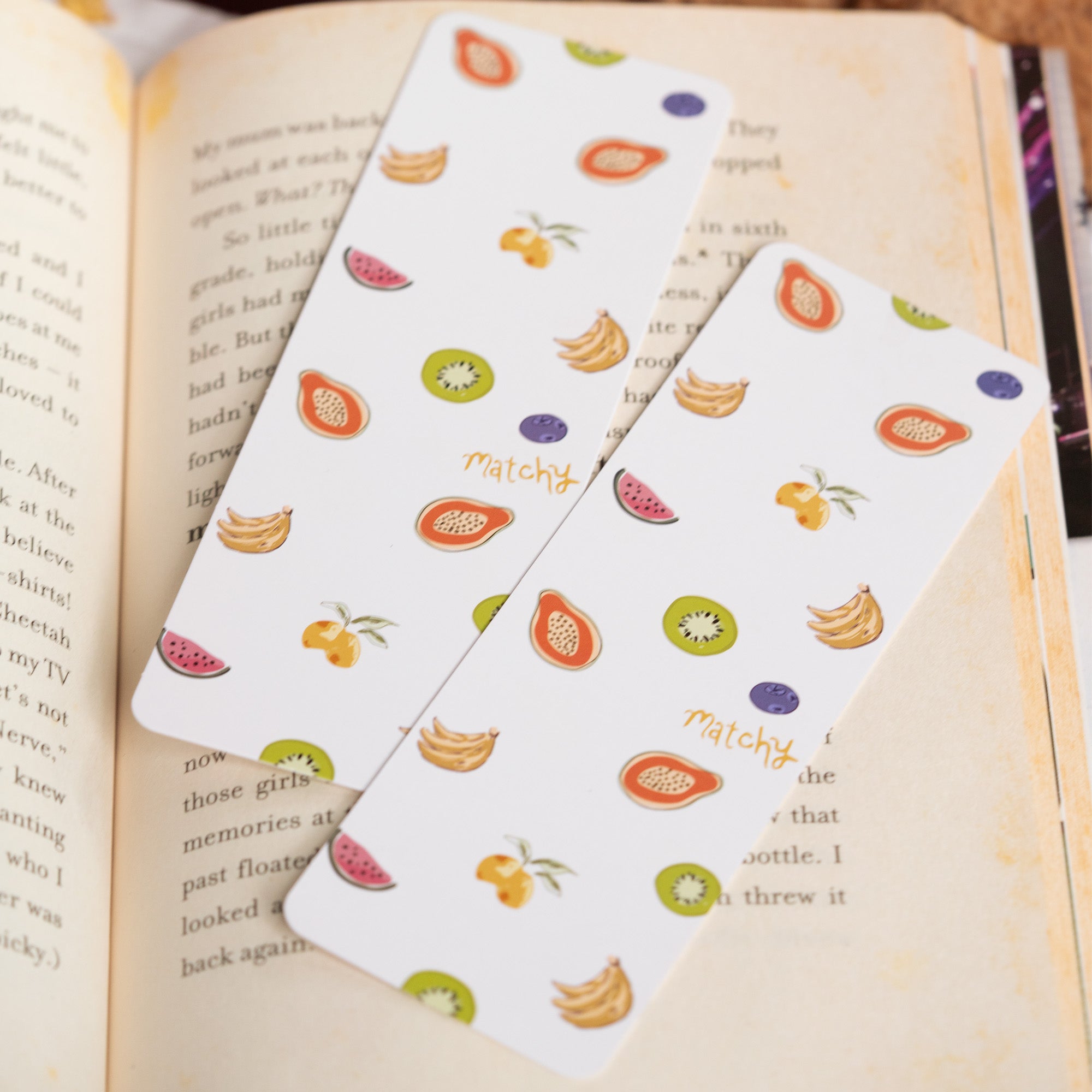Two white bookmarks printed with colourful fruit including kiwi, banana, papaya and watermelon resting on an open book. Matchy Fruity Dayze Bookmark.