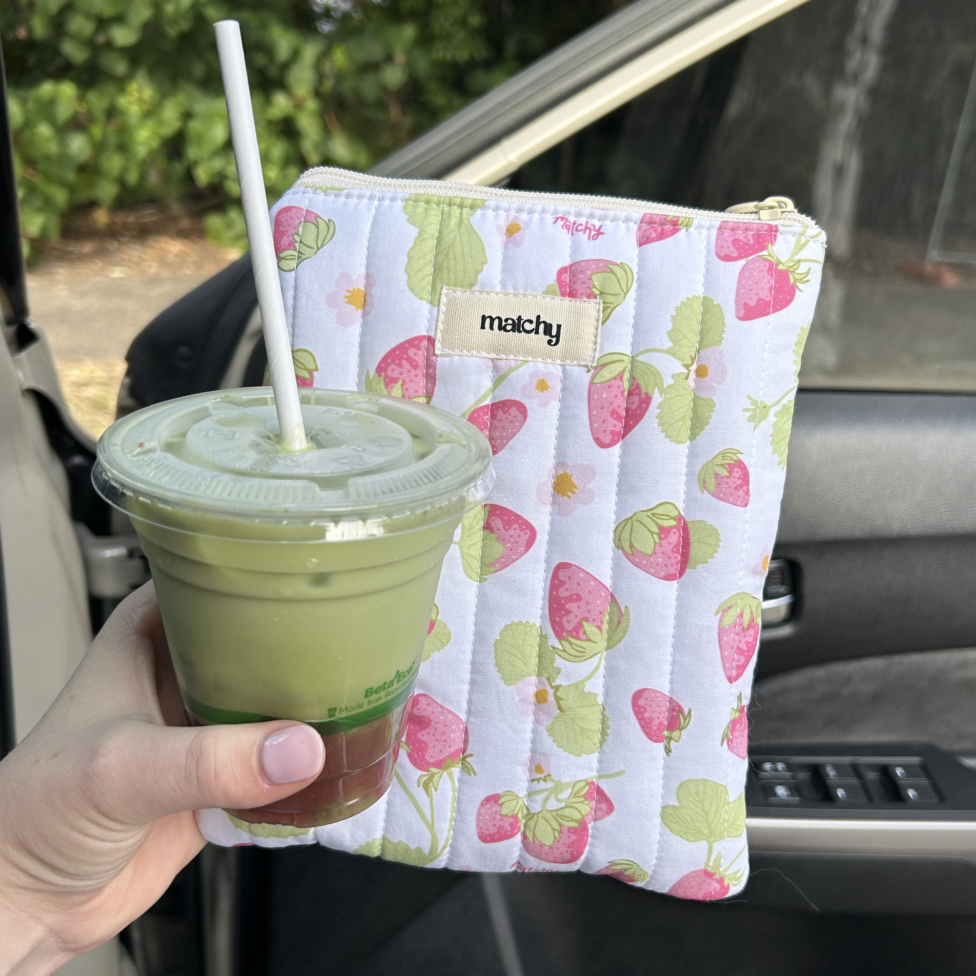 Hand holding a green drink in a cup with a straw, wrapped in a colorful cloth with strawberry pattern, inside a car.