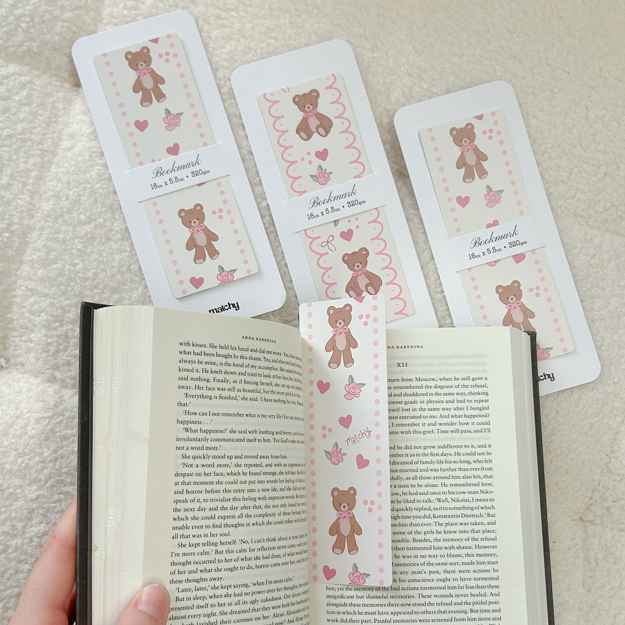 Bookmarks illustrated with brown teddy bears and pink hearts laid on a cream throw, with one tucked into an open book. Matchy Wilbur Bookmark.