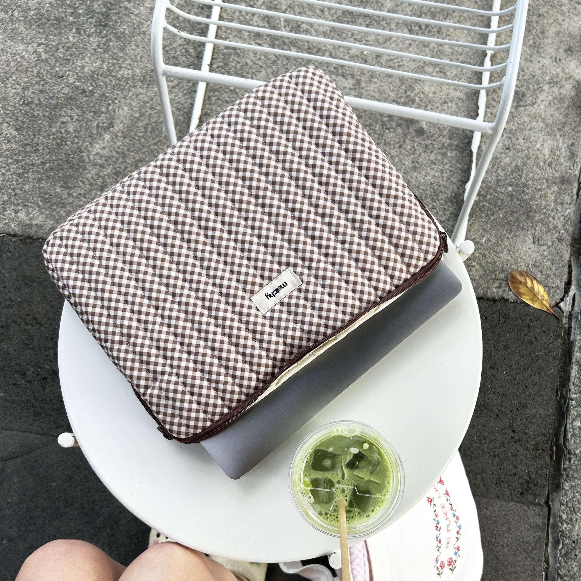 Brown gingham quilted laptop sleeve partly unzipped with a laptop inside, resting on a white cafe table beside an iced matcha. Matchy Georgina Gingham Laptop Sleeve for 13 to 15 inch laptops.