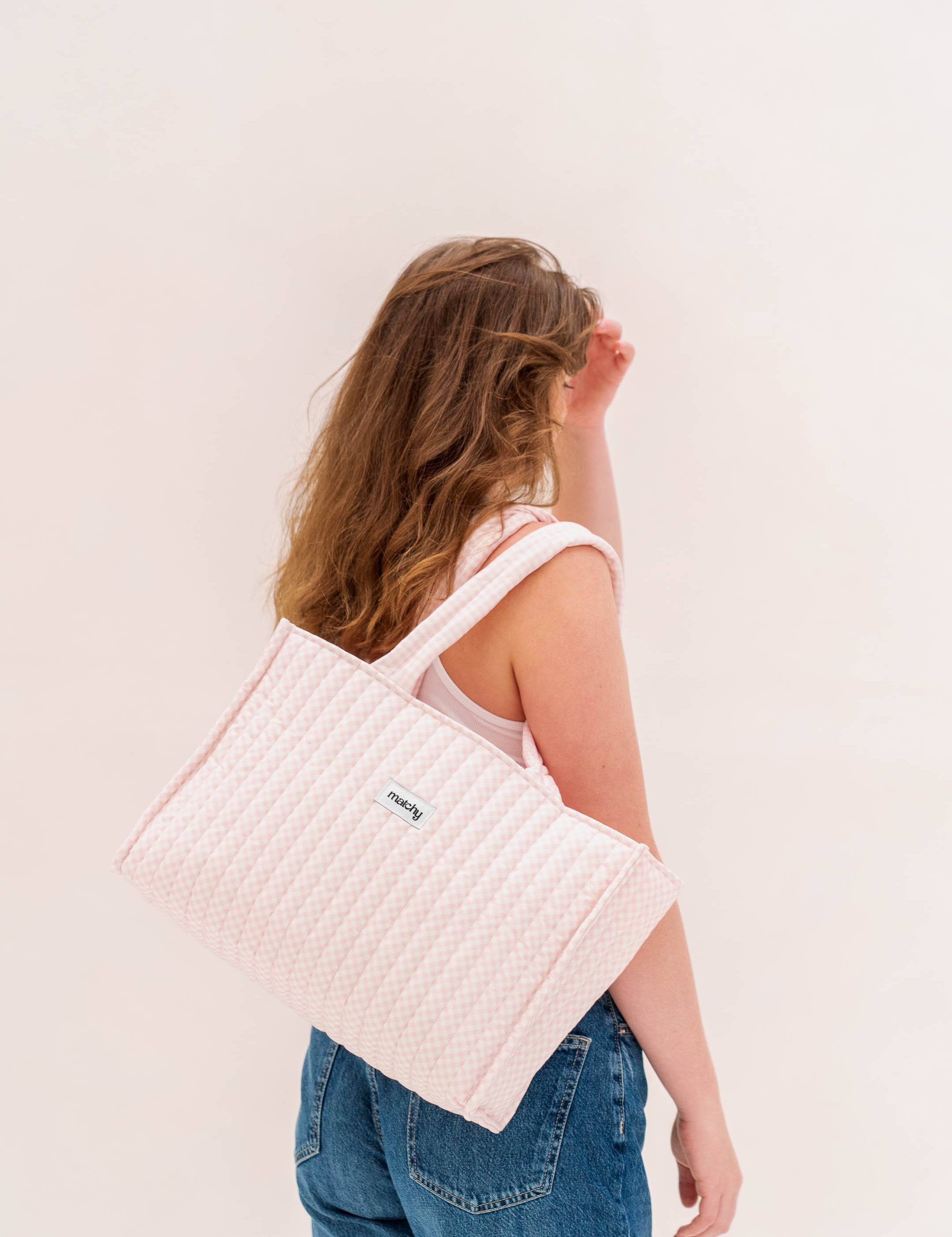 Woman in jeans carrying a soft pink quilted tote bag over her shoulder against a white wall. Matchy Strawberry Take Me With You Tote.