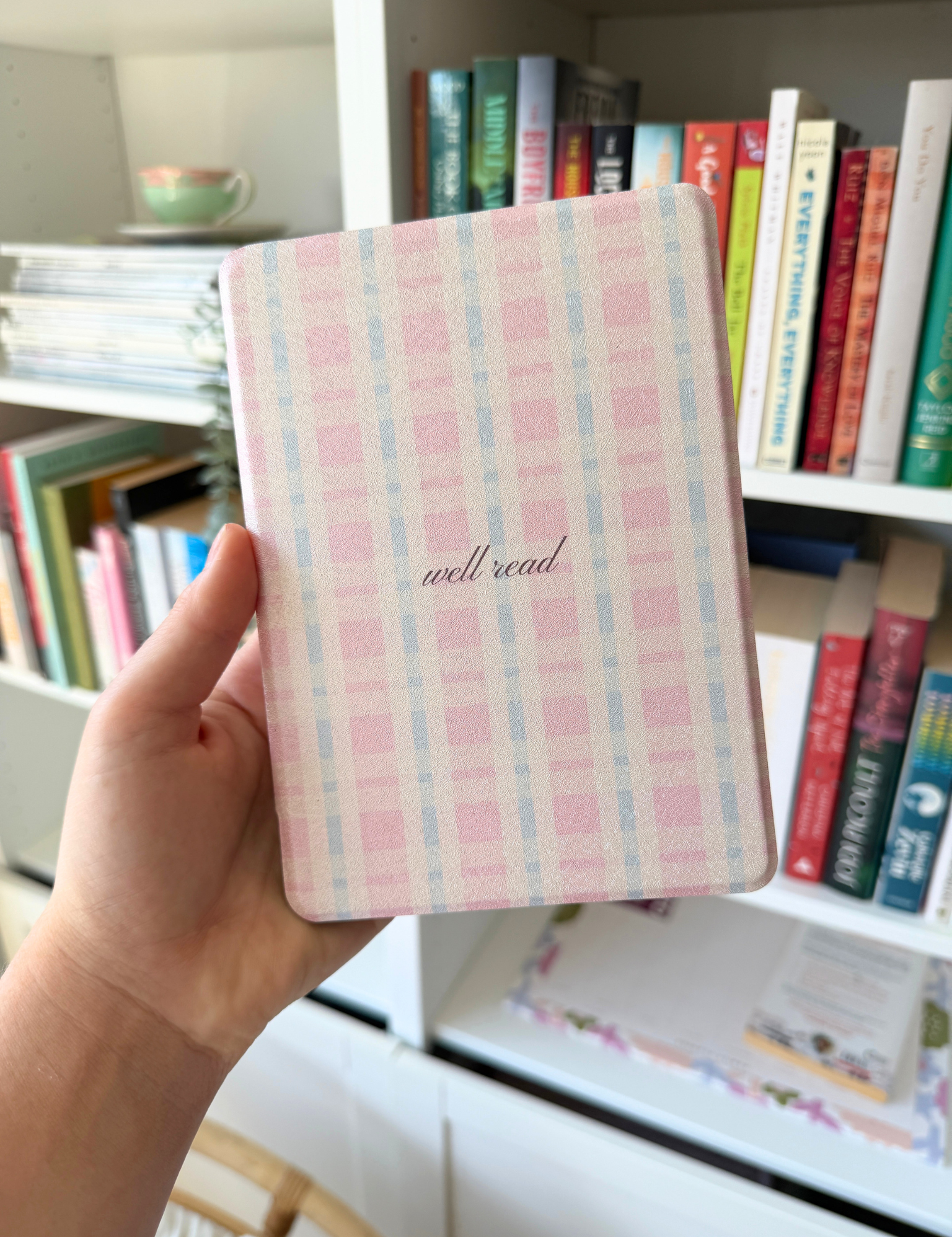 A hand holding a small notebook with a pink and light blue plaid cover and the words 'well read' in cursive, set against a background of a bookshelf filled with books. Kindle Cover with Vegan Leather. Matchy Kindle cover for 6.8 inch Kindle with plastic back.