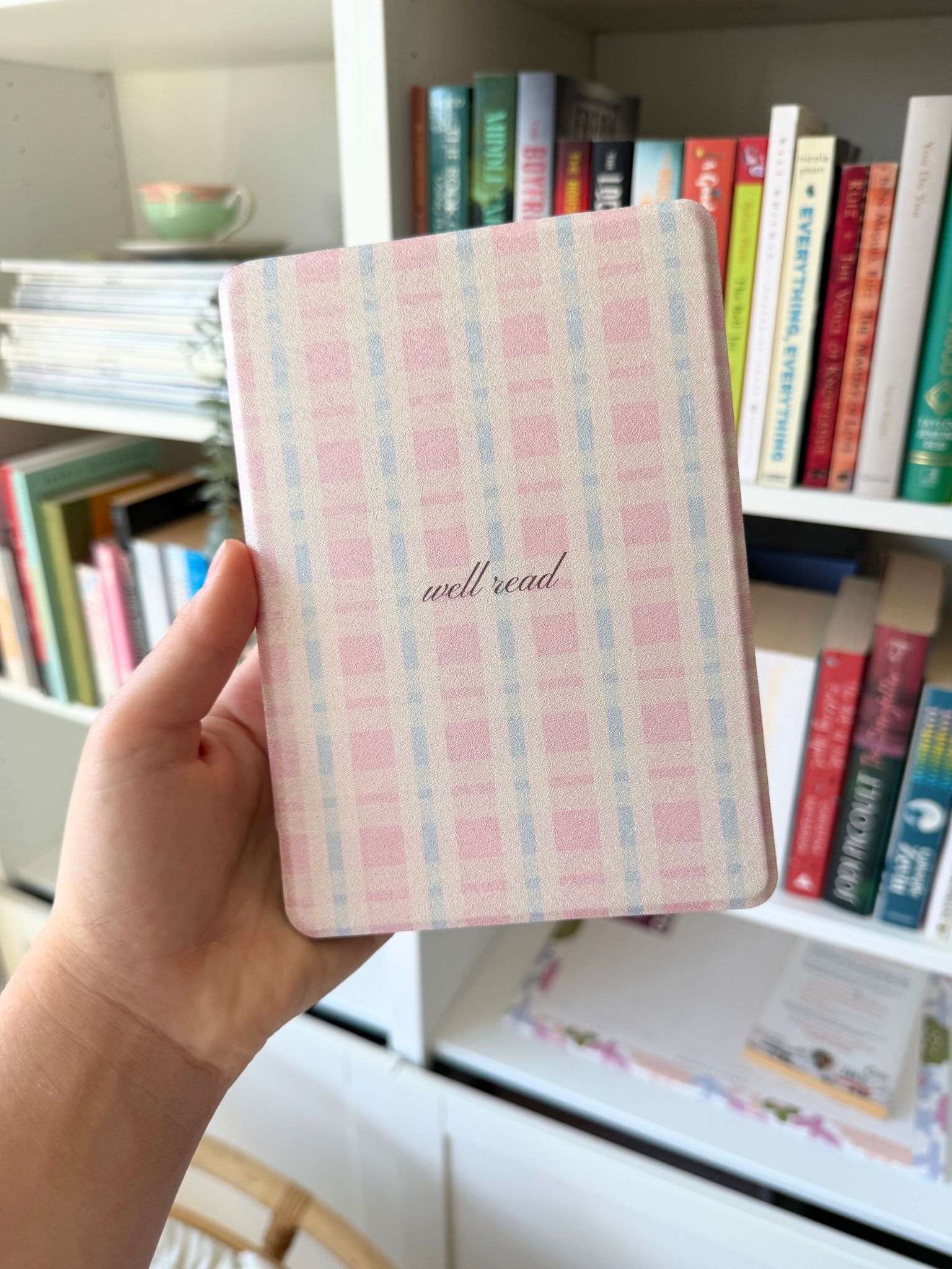A hand holding a small notebook with a pink and light blue plaid cover and the words 'well read' in cursive, set against a background of a bookshelf filled with books. Kindle Cover with Vegan Leather. Matchy Kindle cover for 6.8 inch Kindle with plastic back.