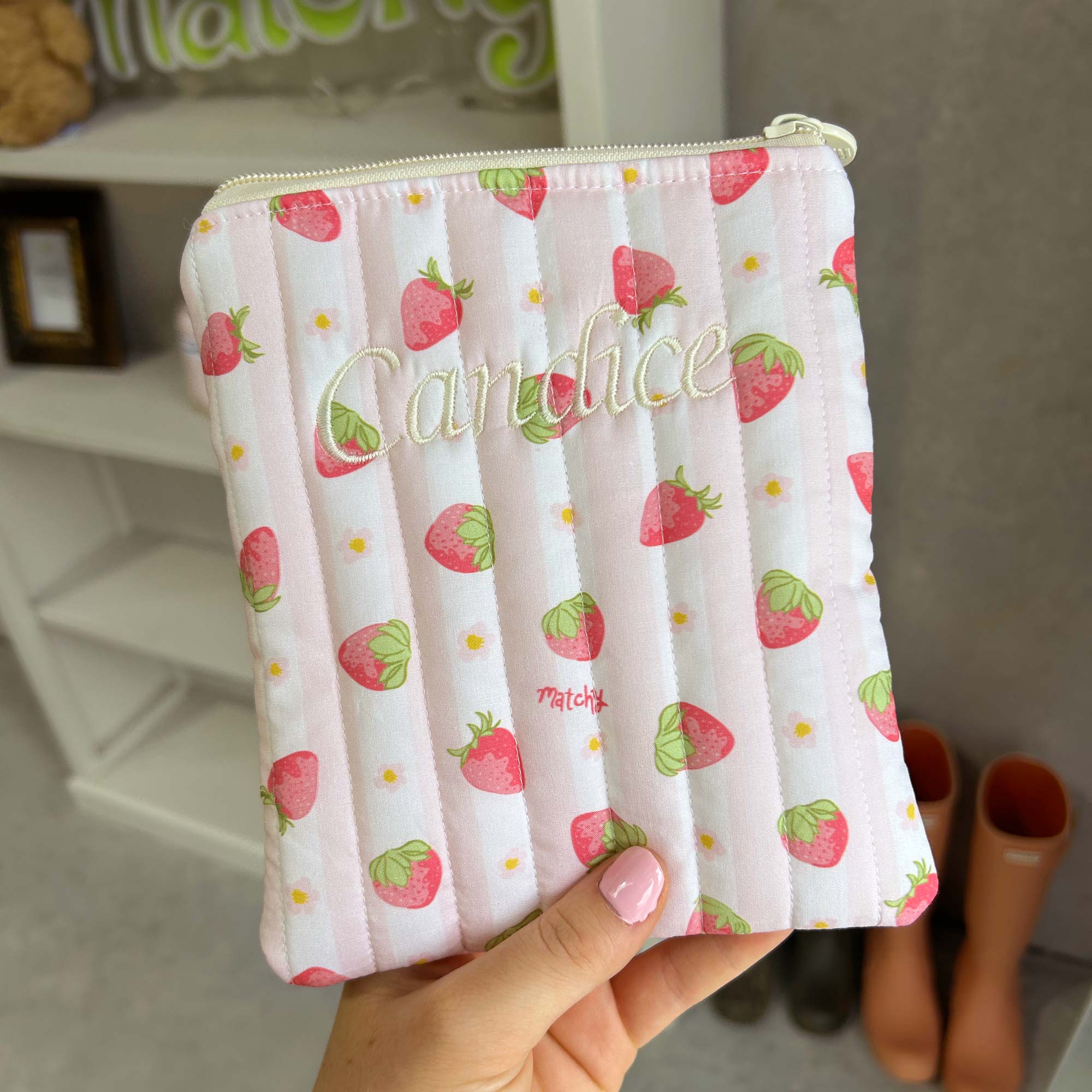 Hand holding a strawberry print quilted eReader sleeve embroidered with a name in cream script. Matchy Sylvie Custom Embroidery eReader Sleeve, a personalised Kindle pouch.