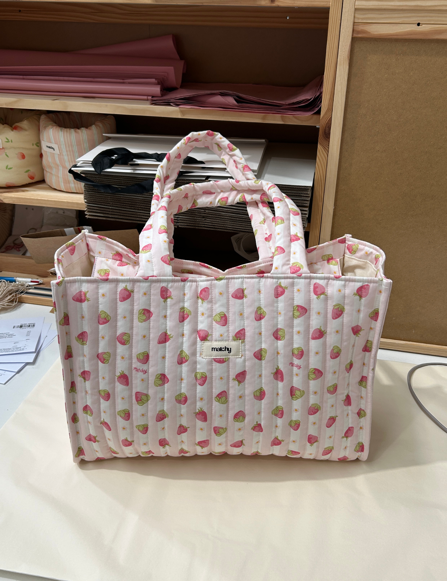 Pale pink strawberry print quilted tote bag standing on a work desk in a studio. Matchy Sylvie Take Me With You Tote.