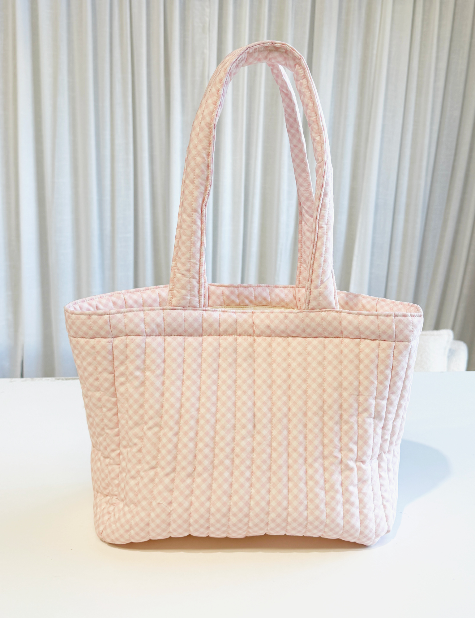 Soft pink gingham quilted tote bag with two shoulder straps standing on a white table. Matchy Wherever You Go Tote in pink, a quilted tote bag.