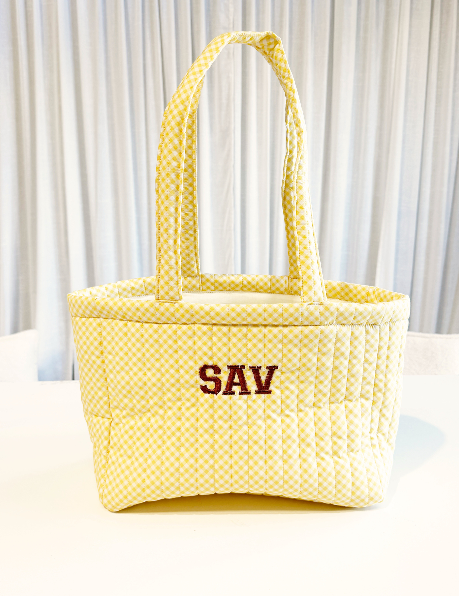 Butter yellow gingham quilted tote bag embroidered with 'SAV' in brown varsity letters, on a white table. Matchy Wherever You Go Tote with custom embroidery.