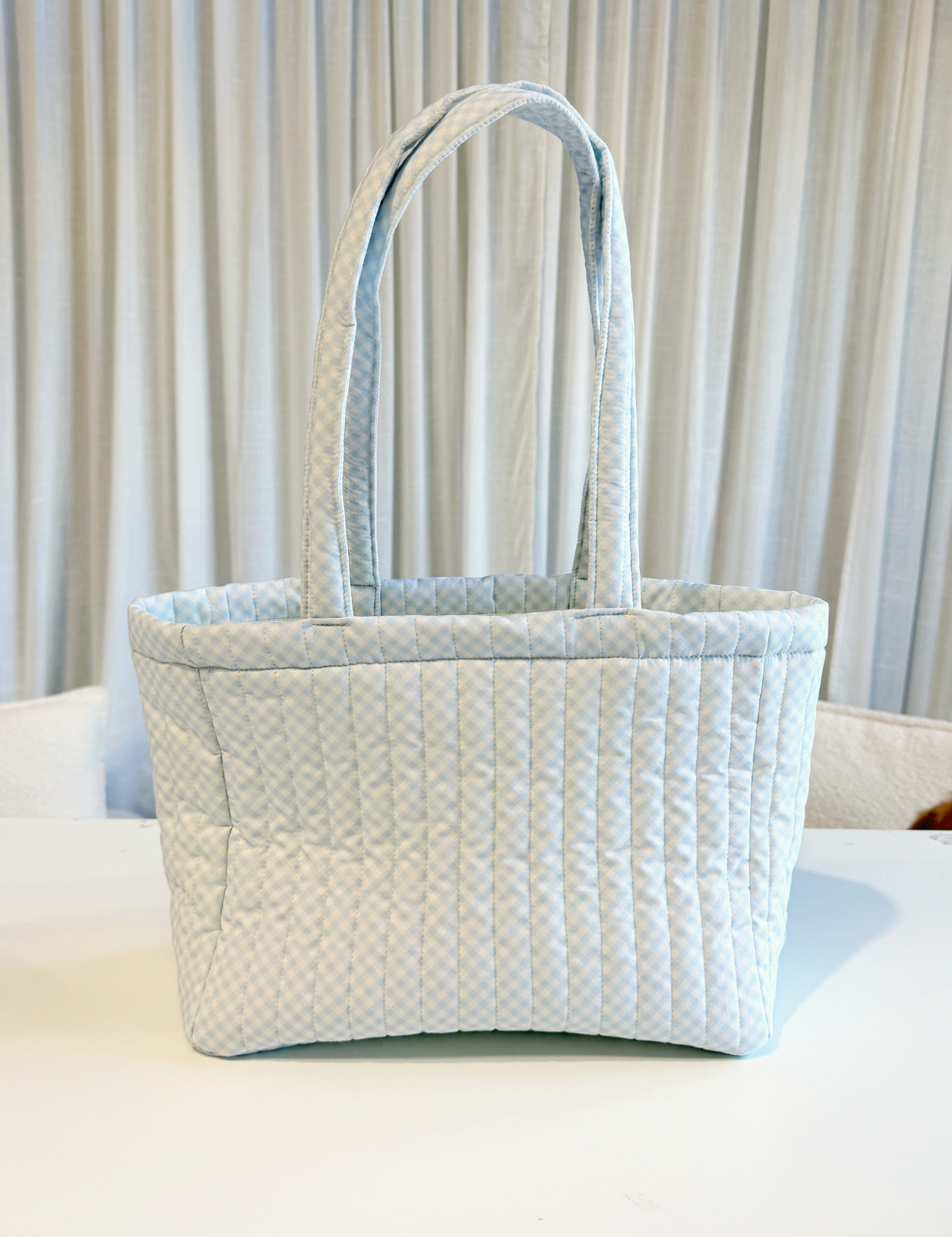 Pale blue gingham quilted tote bag with two shoulder straps standing on a white table. Matchy Wherever You Go Tote in blue, a quilted tote bag.