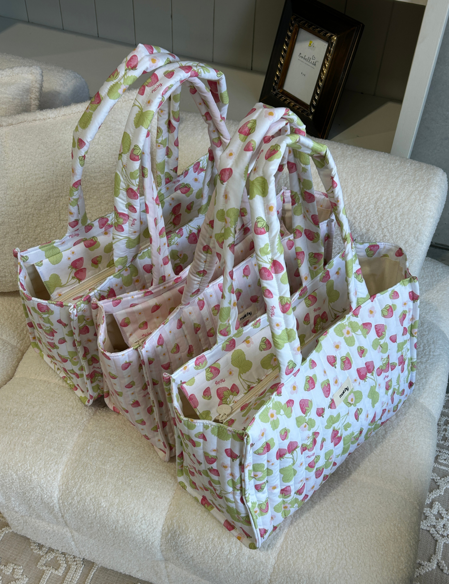 Several strawberry print quilted tote bags lined up on a cream boucle armchair. Matchy Take Me With You Tote collection.