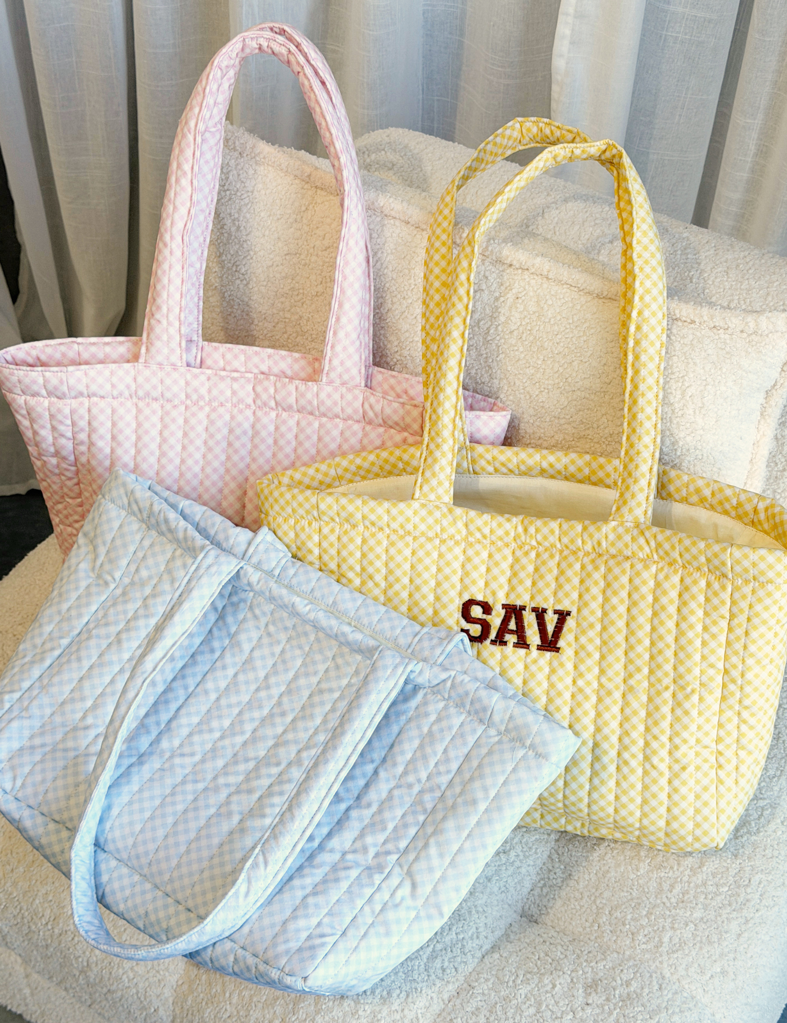 Three quilted tote bags in pink, yellow and blue on a cream chair, the yellow one embroidered with 'SAV' in brown varsity letters. Matchy Wherever You Go Tote, a personalised quilted tote bag.