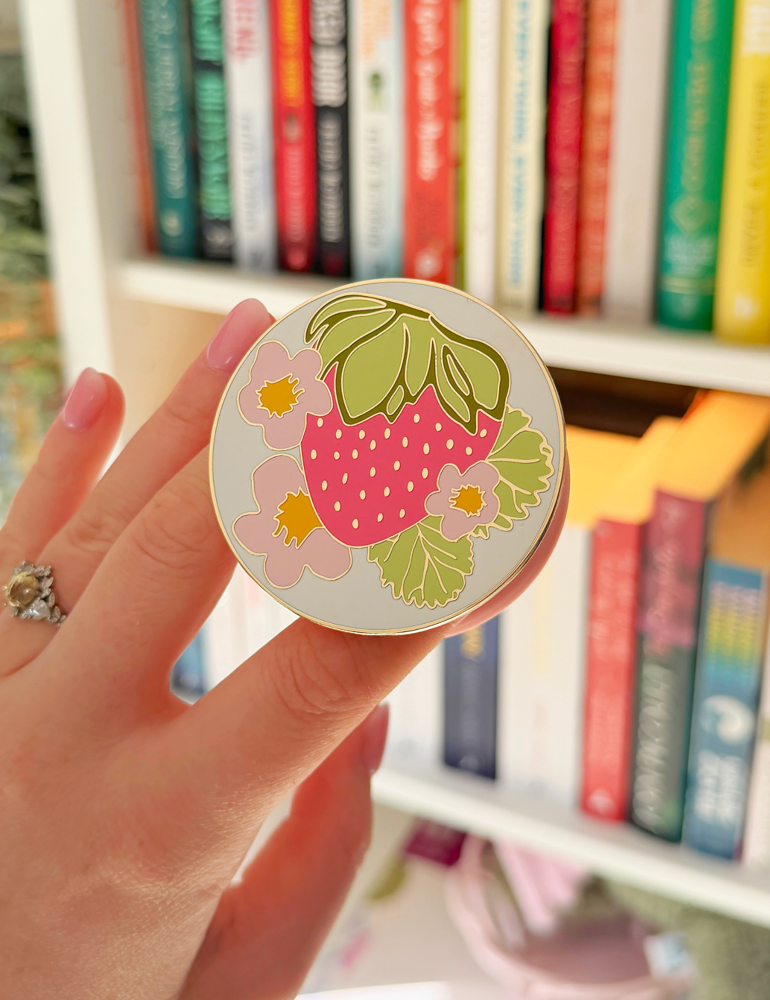 Hand holding a round grip printed with a pink strawberry, green leaves and flowers in front of a bookshelf. Matchy Sofia MagSafe Grip for Kindle and phone.