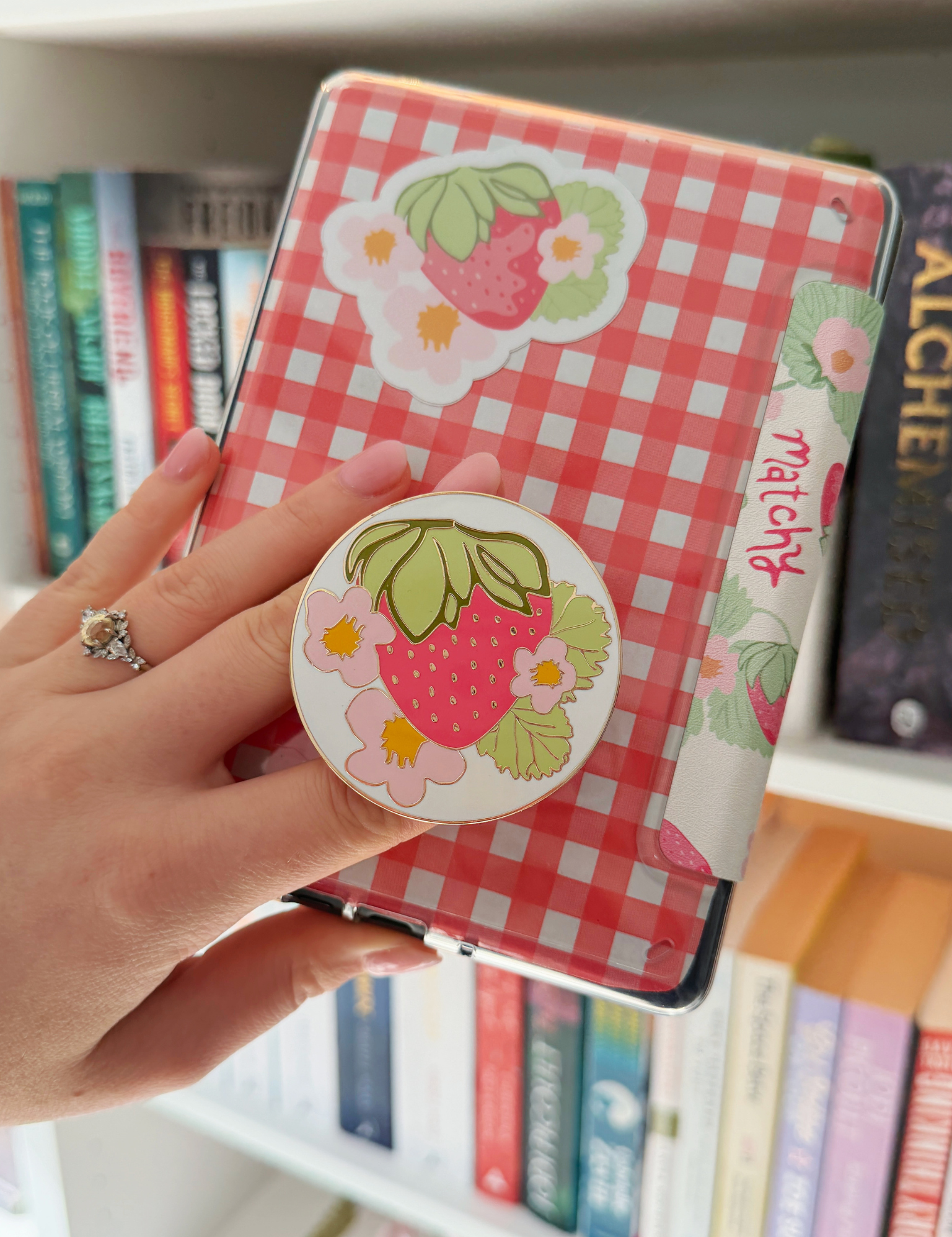 Hand holding a red gingham Kindle case with a strawberry sticker and a round strawberry print grip on the back, in front of a bookshelf. Matchy Sofia MagSafe Grip for Kindle and phone.