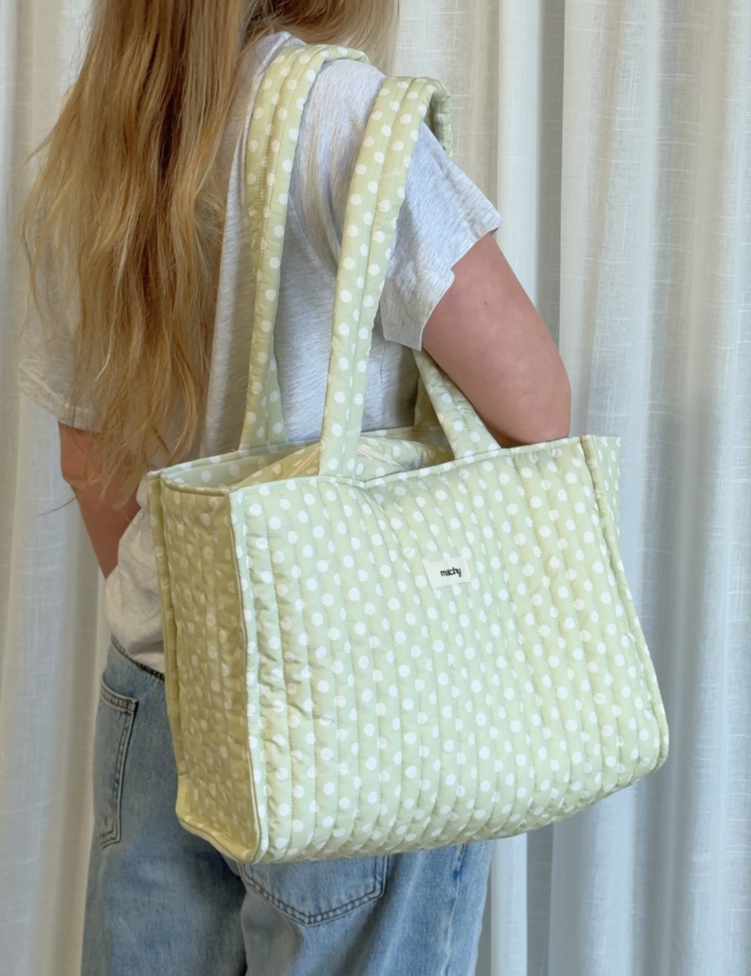 Woman carrying a sage green and white polka dot quilted tote bag over her shoulder. Matchy Poppy Take Me With You Tote, a polka dot quilted tote bag.