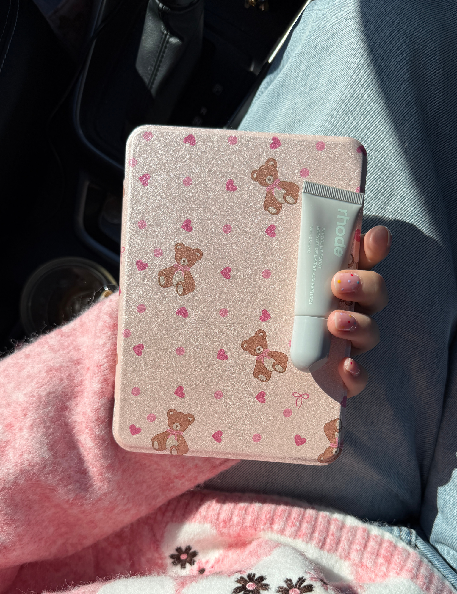 Hand holding a pale pink teddy bear print Kindle case and a tube of lip balm, in a car. Kindle Cover with Vegan Leather. Matchy Kindle cover for 6.8 inch Kindle with plastic back.