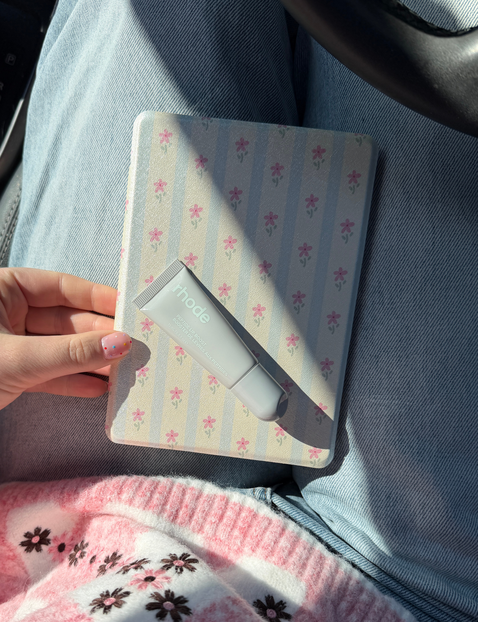 Hand holding a pale blue and cream striped Kindle case with tiny pink flowers and a tube of lip balm, in a car. Kindle Cover with Vegan Leather. Matchy Kindle cover for 6.8 inch Kindle with plastic back.