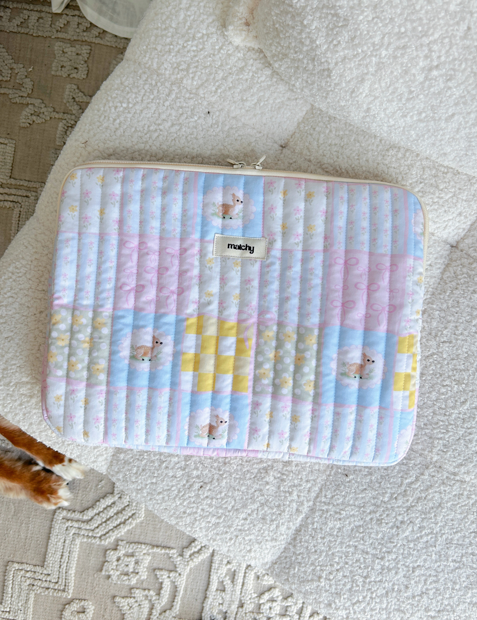 Pastel patchwork quilted laptop sleeve in pink, blue and yellow squares with deer motifs, lying on a cream textured rug. Matchy Patchwork Laptop Sleeve for 13 to 15 inch laptops.