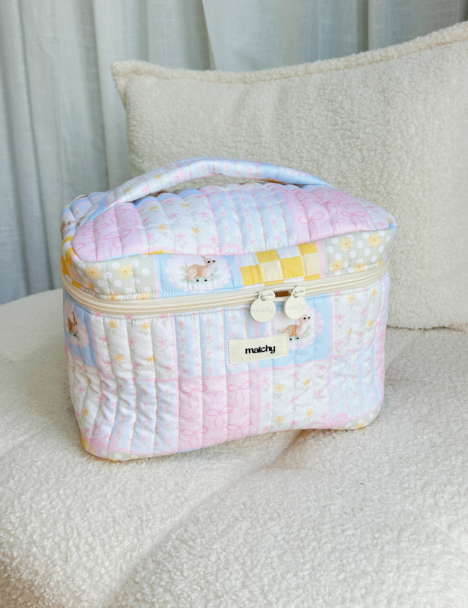Pastel patchwork quilted train case with a deer motif and cream zip sitting on a cream boucle chair. Matchy Patchwork Train Case, a quilted makeup and toiletry case.