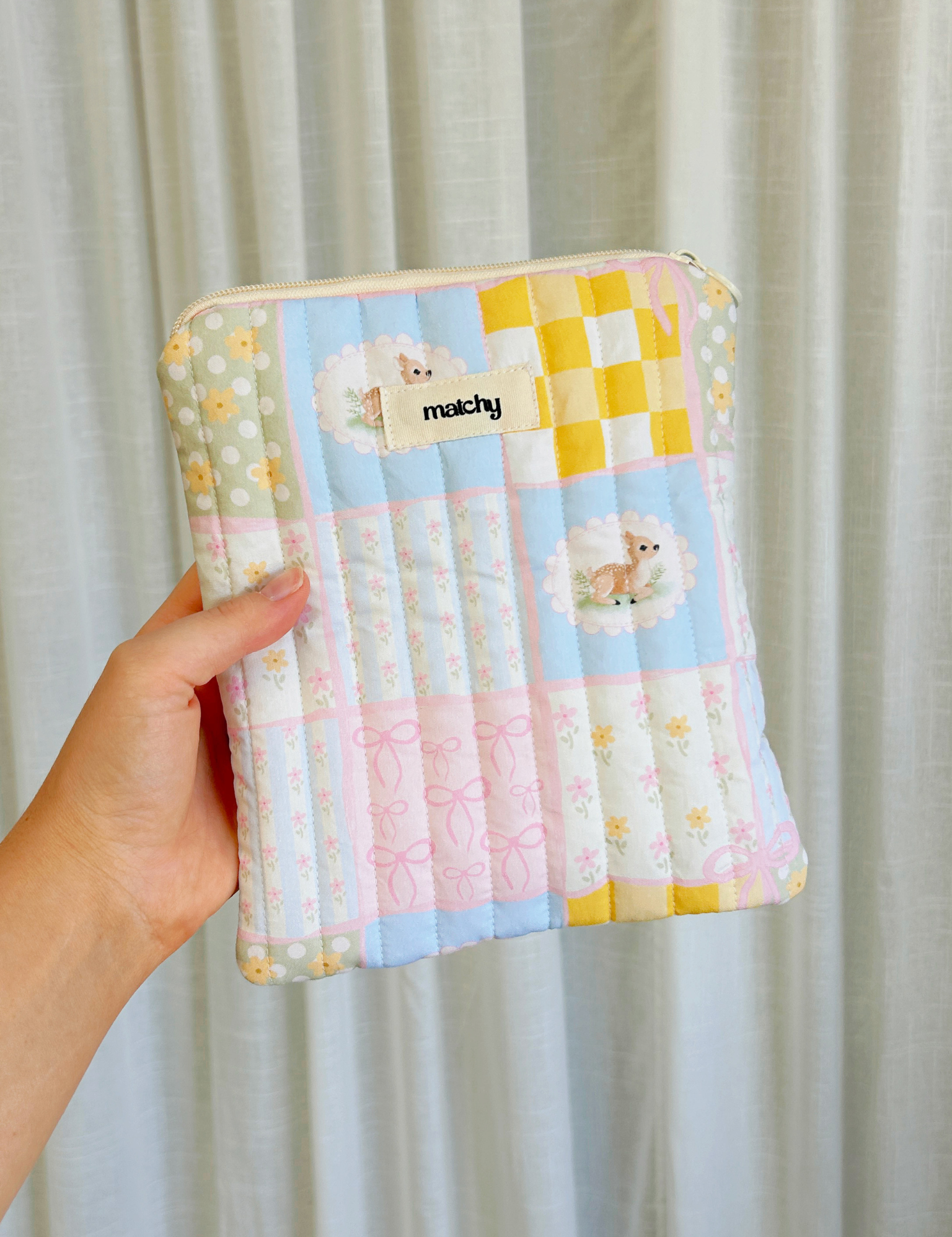 Hand holding a pastel patchwork quilted eReader sleeve in pink, blue and yellow squares with deer motifs. Matchy Patchwork eReader Sleeve, a padded Kindle pouch.
