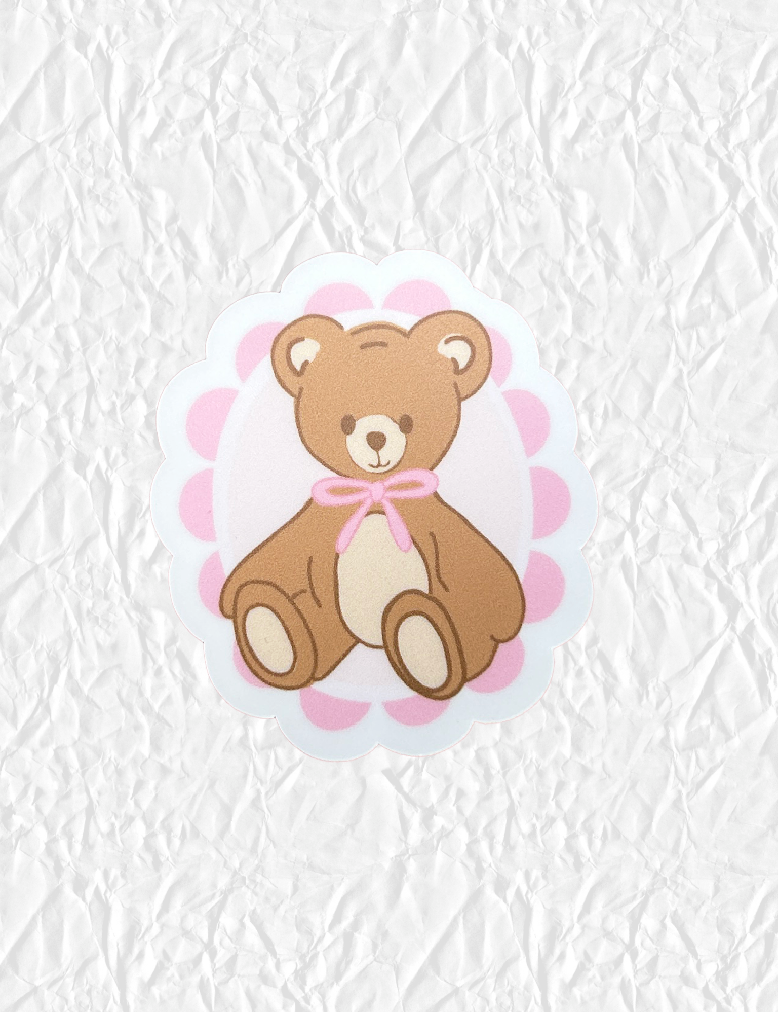Teddy bear sticker with a pink bow shown on crumpled white paper. Matchy Teddy Valentine Sticker.