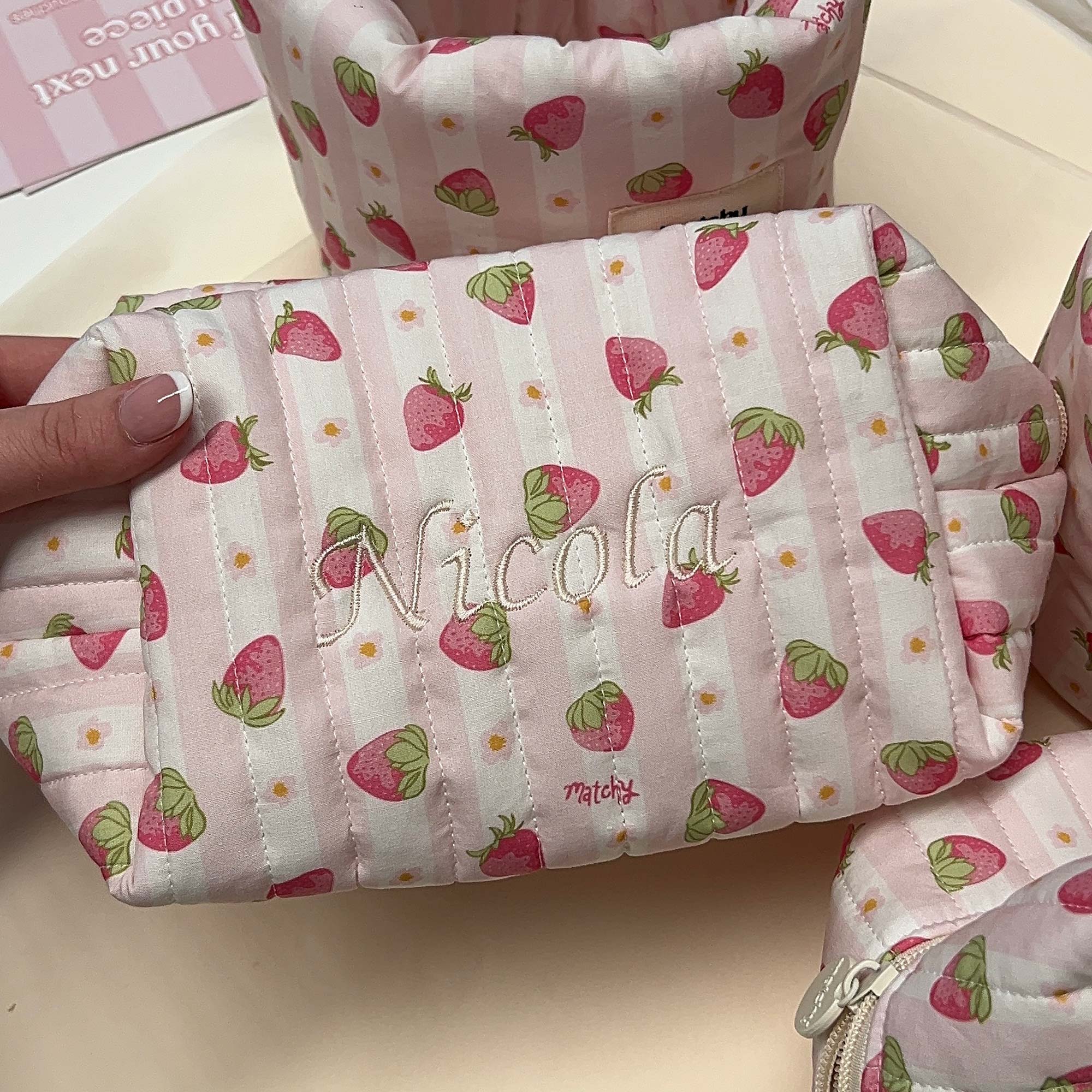 Pale pink strawberry print quilted carry-all bag embroidered with a name in cream script, surrounded by matching bags. Matchy Sylvie Custom Embroidery Carry-All Bag.