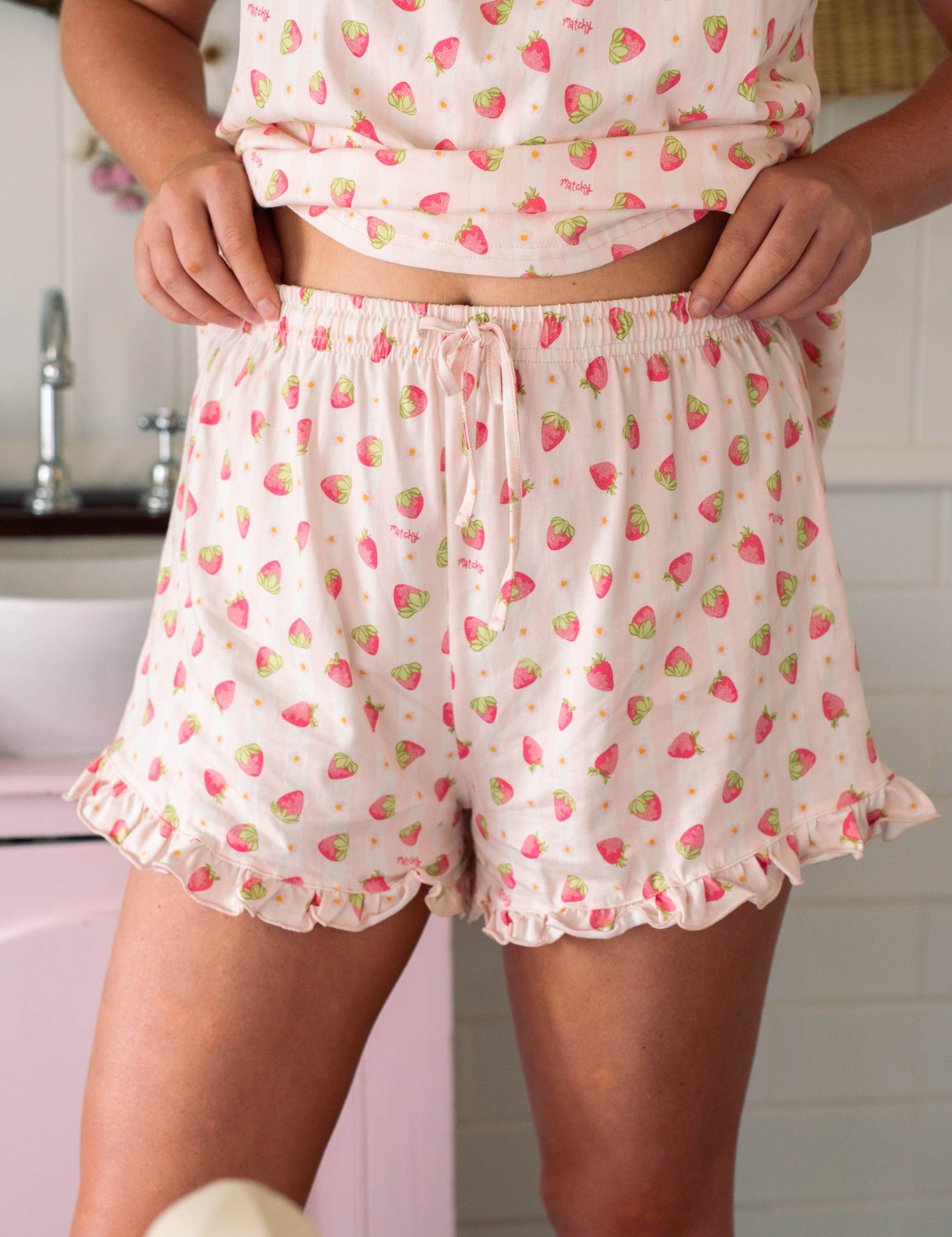 Front view of white sleep shorts with a small pink strawberry print, drawstring waist and ruffle hems. Matchy Sylvie Ruffle Short, cotton pyjama shorts.